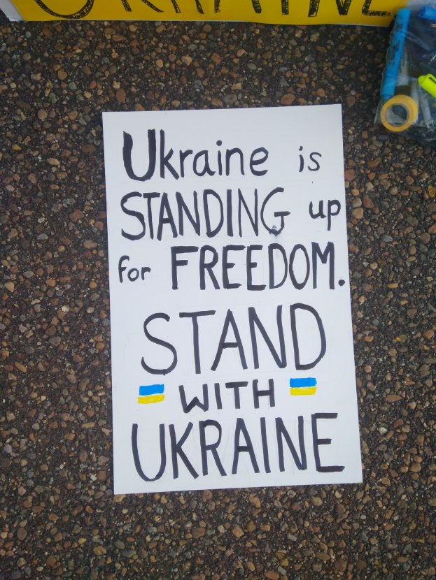 Ein weißes Blatt Papier mit dem Text "Ukraine steht für Freiheit auf, steht mit Ukraine" vor einem gelben Banner auf dem Boden liegend, mit einer Plastikabdeckung mit verschiedenen Gegenständen rechts daneben.