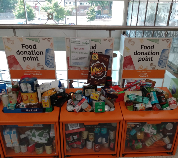 Orange Schränke in einem Gebäude mit gespendeten Lebensmitteln wie Flaschen, Dosen und Kartons, mit Informationsschildern und einem Blick auf Fahrzeuge, Bäume, Gebäude und Himmel durch Glasfenster.