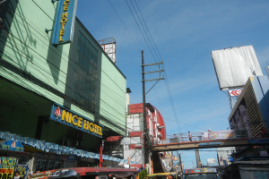 Eine belebte Stadtstraße mit Fahrzeugen, Gebäuden, Strommasten, einer Brücke mit Fußgängern und einem bewölkten Himmel.