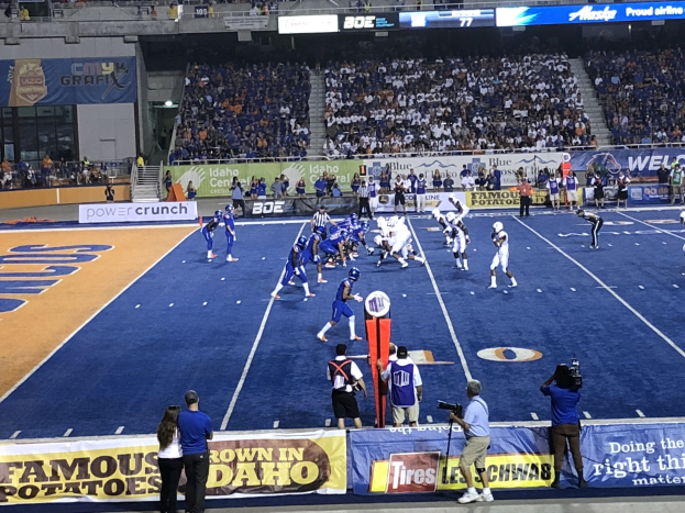 Ein Fußballspiel in einem Stadion mit Zuschauern, die stehen und sitzen, Schilder, eine Person mit einer Kamera auf einem Stativ und verschiedene Gegenstände im Hintergrund, einschließlich Geländern, Treppen und Tafeln mit Text.