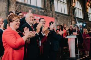 Eine Gruppe von Menschen, wahrscheinlich Liberale, steht vor einer Menge und klatscht feierlich, mit einem Podium, Mikrofon und Texttafel rechts, sowie Stühlen, einem Banner, Wand, Fenstern und Lichtern im Hintergrund.