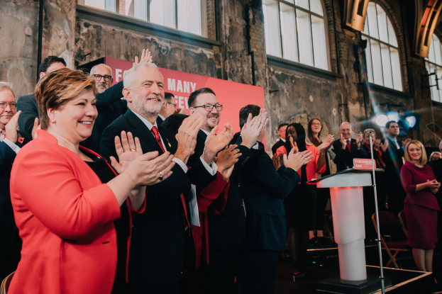 Eine Gruppe von Menschen, wahrscheinlich Liberale, steht vor einer Menge und klatscht feierlich, mit einem Podium, Mikrofon und Texttafel rechts, sowie Stühlen, einem Banner, Wand, Fenstern und Lichtern im Hintergrund.
