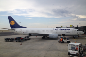 Ein Lufthansa Airbus A320-200-Flugzeug steht auf dem Frankfurter Flughafen, umgeben von verschiedenen Bodenfahrzeugen, Wagen, Absperrgittern, Pfosten und Texttafeln unter einem bewölkten Himmel.