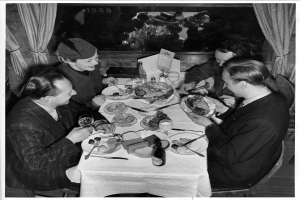 Eine Gruppe von Menschen sitzt um einen Tisch mit Tellern, Gläsern, Flaschen und Besteck, mit Vorhängen im Hintergrund.