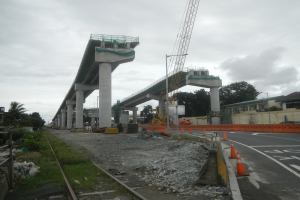 Eine Baustelle mit einer Brücke im Hintergrund, eine Straße mit Absperrkegeln auf der rechten Seite, Steine und Gras am Boden, eine Eisenbahnschiene auf der linken Seite, Bäume und Gebäude auf beiden Seiten der Straße und ein bewölktes Himmel.