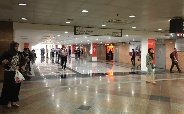 Menschen mit Taschen durch eine Flughafenhalle gehen, mit Texttafeln an den Wänden, Deckenbeleuchtung und einem Mülleimer im Hintergrund; eine Frau ist in Schuhen zu sehen.