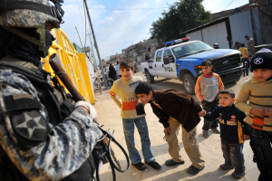 Eine Gruppe von Kindern, die vor einem Polizisten mit einer Waffe steht, mit Fahrzeugen, Menschen, Fahrrädern und Bäumen im Hintergrund unter einem bewölkten Himmel.