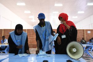Eine Gruppe von Menschen steht um einen Tisch mit einem Megafon, Papieren und anderen Gegenständen in einer Wahllokal mit mehr Menschen im Hintergrund, unter einer Decke mit Lampen.