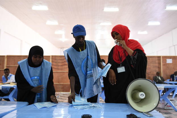 Eine Gruppe von Menschen steht um einen Tisch mit einem Megafon, Papieren und anderen Gegenständen in einer Wahllokal mit mehr Menschen im Hintergrund, unter einer Decke mit Lampen.