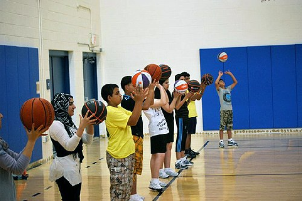 Gruppe junger Menschen mit Basketballs auf einem Basketballfeld während eines Camps, mit Türen und einer Wand im Hintergrund.