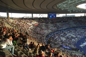 Eine große Menschenmenge sitzt in einem Stadion und schaut ein Fußballspiel, mit einer Bühne rechts, Fahnen, Stangen und einem Bildschirm im Hintergrund, unter einem sichtbaren Himmel, im Allianz Stadion in München, Deutschland.