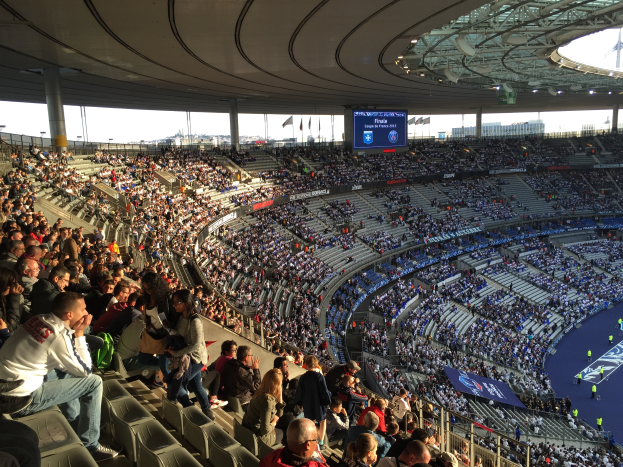 Eine große Menschenmenge sitzt in einem Stadion und schaut ein Fußballspiel, mit einer Bühne rechts, Fahnen, Stangen und einem Bildschirm im Hintergrund, unter einem sichtbaren Himmel, im Allianz Stadion in München, Deutschland.