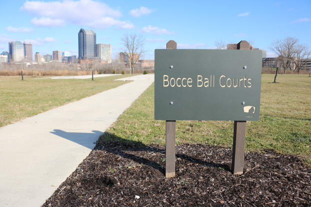 Ein Schild am Boden in der Nähe eines Weges und Bäume mit der Aufschrift "Bocce Ball Courts in Indianapolis, Indiana" und einem bewölkten Himmel darüber.