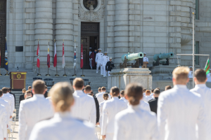 Gruppe von Menschen in weißen Marineuniformen vor einem Gebäude mit Säulen und Treppe während einer Abschlussfeier mit Fahnen, Podium und Kanonen im Hintergrund.