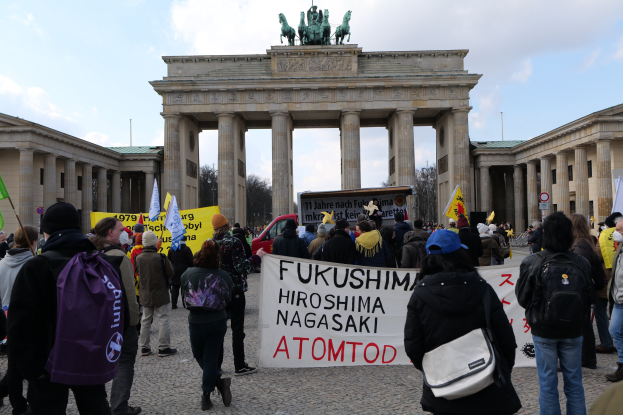 Demonstranten mit Schildern und Fahnen mit der Aufschrift 'Fukushima Hiroshima Nagasaki Atomod' vor dem Brandenburger Tor in Berlin, mit den Säulen und Statuen des Tors im Hintergrund.