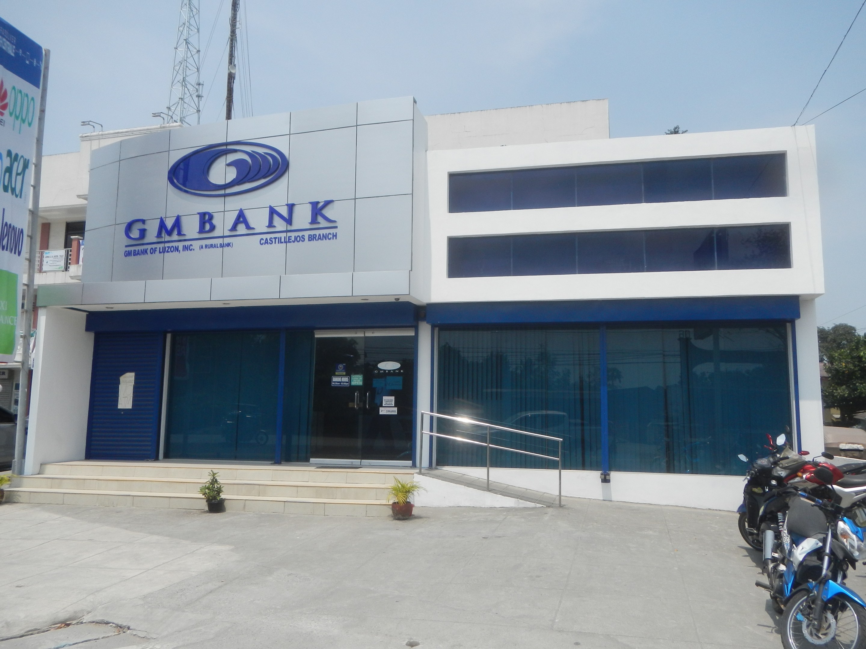Außenansicht der Gmbank Bank in Hyderabad mit Glaswänden, Treppen mit Geländern, Topfpflanzen, Straße mit Fahrzeugen, einem Banner, einem Turm mit Drähten, Bäumen und einem bewölkten Himmel.
