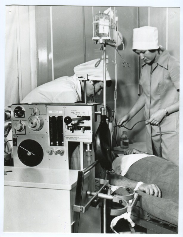 Schwarz-weiß-Foto von zwei Schwestern in einem Operationssaal, eine liegt auf einem Bett und die andere steht, waßrend sie ein Objekt hält, mit medizinischer Ausrüstung im Hintergrund.