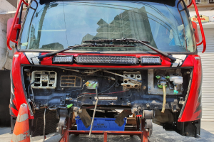 Ein roter Lastwagen steht vor einem Geb\c3ude mit einem Verkehrskegel links daneben und einem Rollladen rechts daneben, w\c3hrde Schilder mit Text und Zahlen im Hintergrund sichtbar sind.