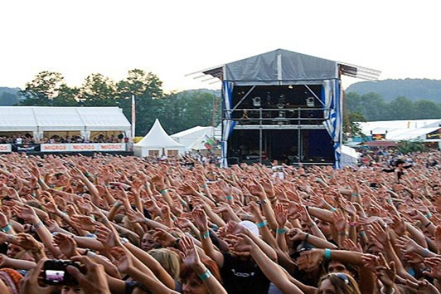 Eine große Menschenmenge auf einem Musikfestival mit erhobenen Händen, einige halten Telefone, vor einer Bühne mit Performern, weißen Zelten, Bäumen und einem klaren blauen Himmel.