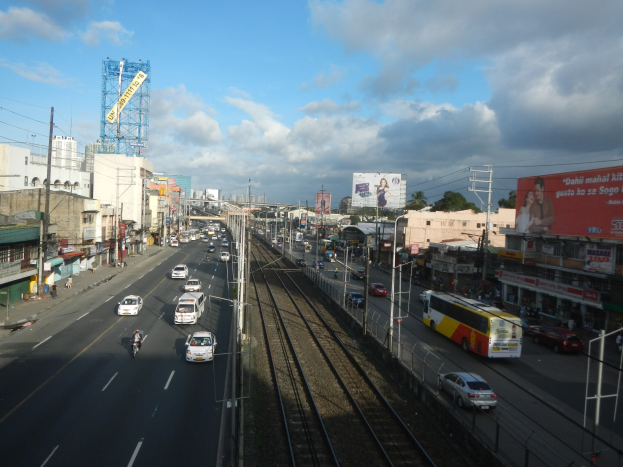 Eine belebte Stadtstraße mit Fahrzeugen, Strommasten, Gebäuden, Schildern, Bäumen und einem bewölkten Himmel neben einer Bahnschiene.