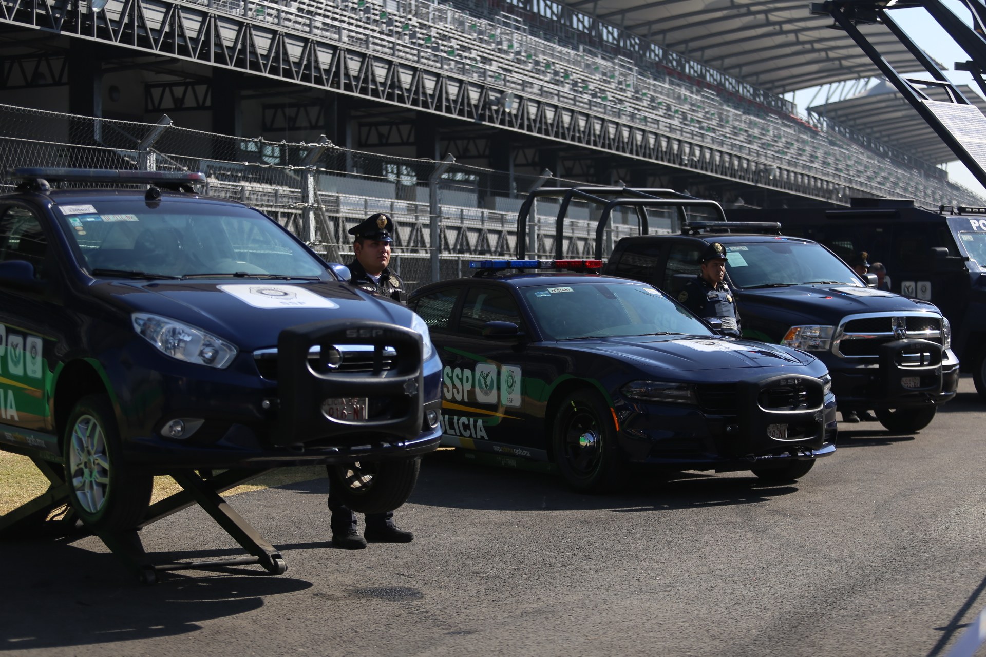 Polizeiautos vor einer Rennstrecke mit uniformierten Beamten auf der Straße, Stadion und Absperrungen im Hintergrund.