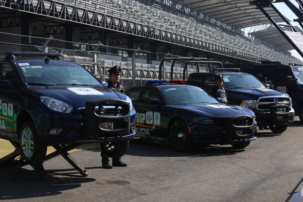 Polizeiautos vor einer Rennstrecke mit uniformierten Beamten auf der Straße, Stadion und Absperrungen im Hintergrund.