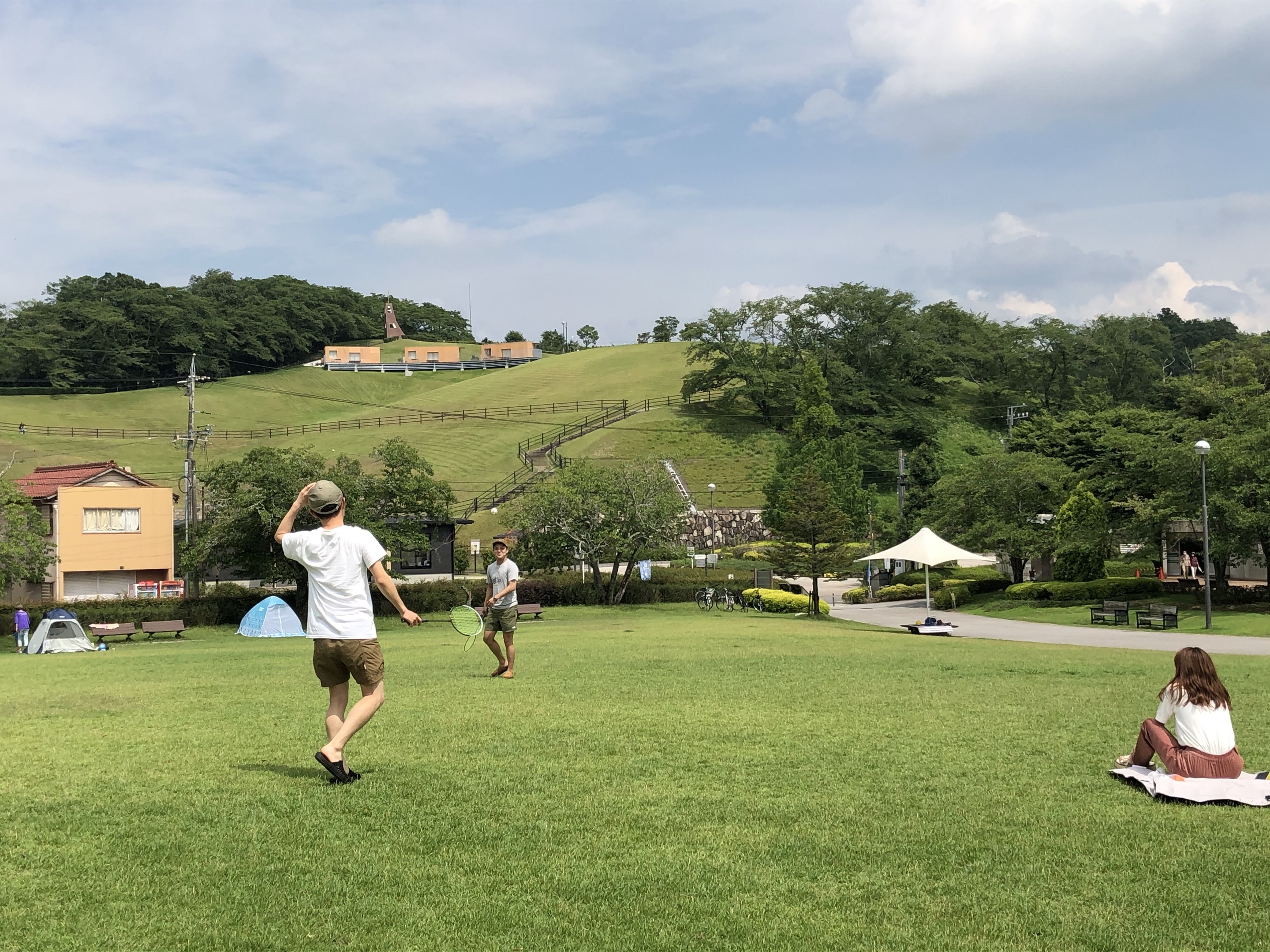 Eine Gruppe von Menschen, die Badminton in einem Park spielt, mit einem Mann, der einen Schläger hält, anderen, die auf dem Gras sitzen, und Zelten, Straßenlaternen, Gebäuden, Bäumen, Hügeln und Wolken im Hintergrund.