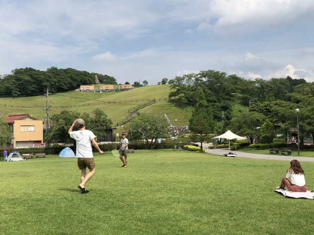 Eine Gruppe von Menschen, die Badminton in einem Park spielt, mit einem Mann, der einen Schläger hält, anderen, die auf dem Gras sitzen, und Zelten, Straßenlaternen, Gebäuden, Bäumen, Hügeln und Wolken im Hintergrund.