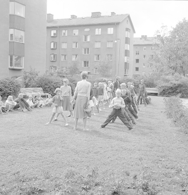 Schwarzes Foto von Kindern beim Fussballspielen in einem Park mit Gebäuden, Bäumen, Pflanzen und Bänken im Hintergrund.