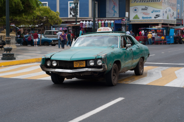 Ein grünes Auto fährt eine Straße entlang, gesäumt von hohen Gebäuden, Fußgängern auf dem Gehweg, Laternenmasten und Bäumen, mit Ständen und Schildern im Hintergrund, die Text und Bilder anzeigen.