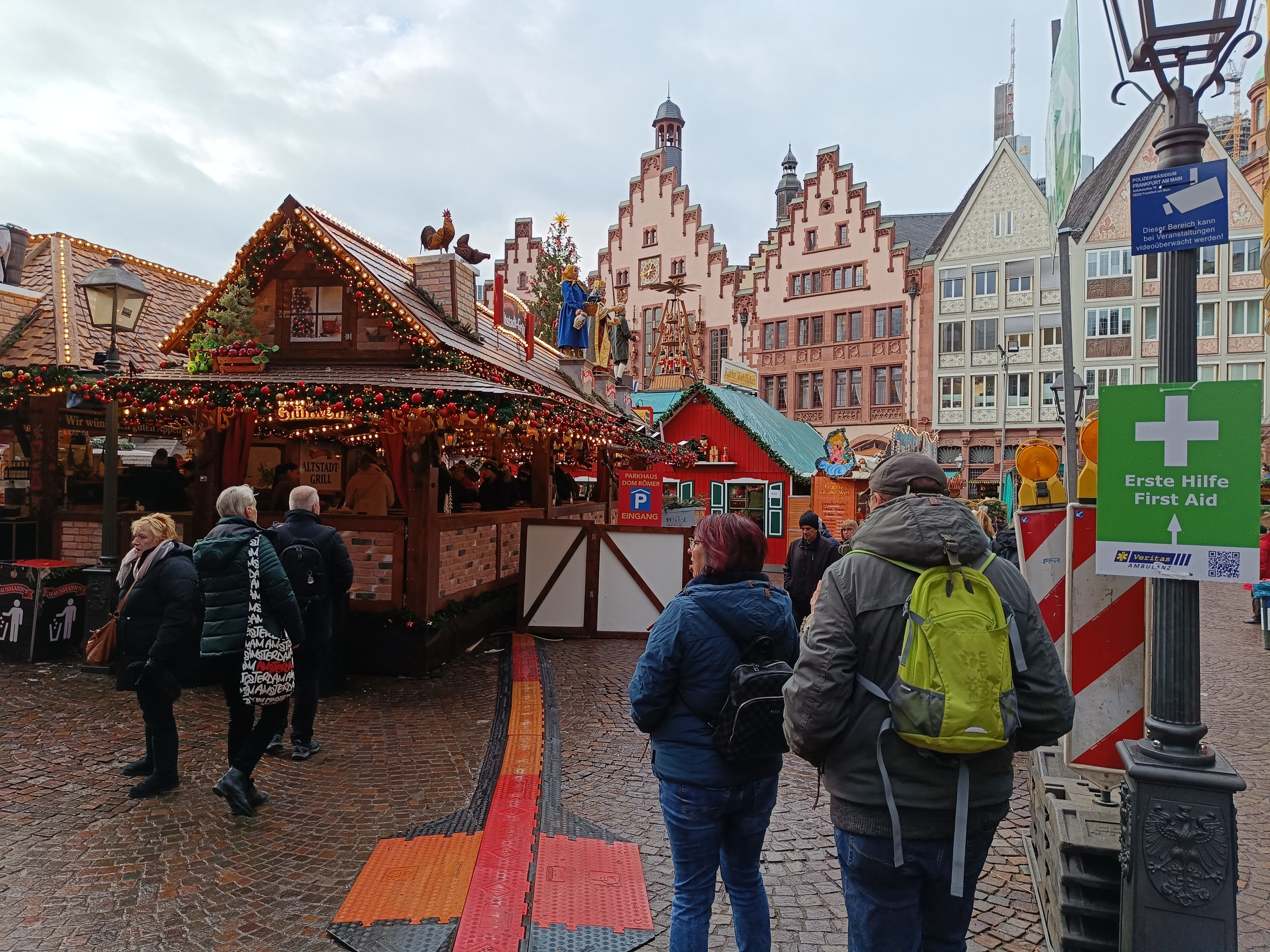 Menschen auf einem Kopfsteinpflasterweg neben einem Weihnachtsmarkt in Nürnberg, Deutschland, mit Laternenmasten, Informationsschildern und Gebäuden im Hintergrund.