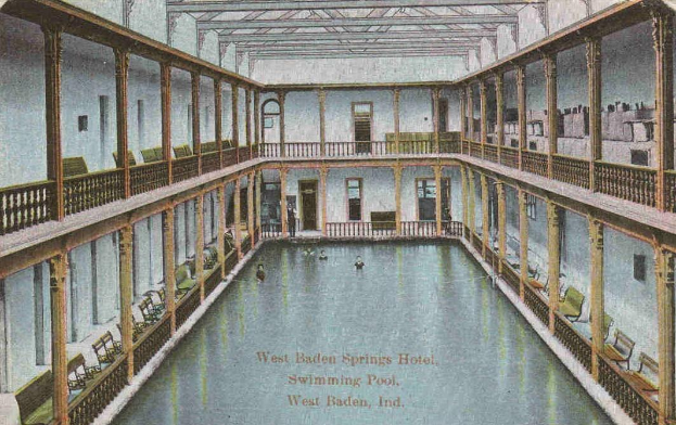 Eine alte Postkarte zeigt ein Hallenbad im West Baden Springs Hotel mit Menschen im Wasser und verschiedenen architektonischen Elementen drumherum sowie Text am unteren Rand.