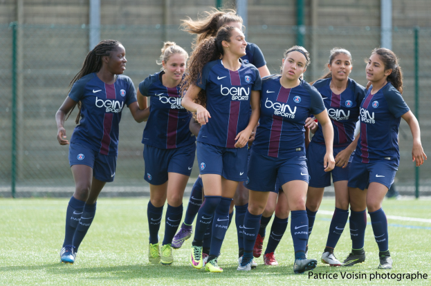 Eine Gruppe junger Frauen beim Fußballspielen auf einem Rasenplatz mit Maschendrahtzaun und einer Wand im Hintergrund, mit dem Text "Paris Saint-Germain Frauenfußball" in der rechten unteren Ecke.
