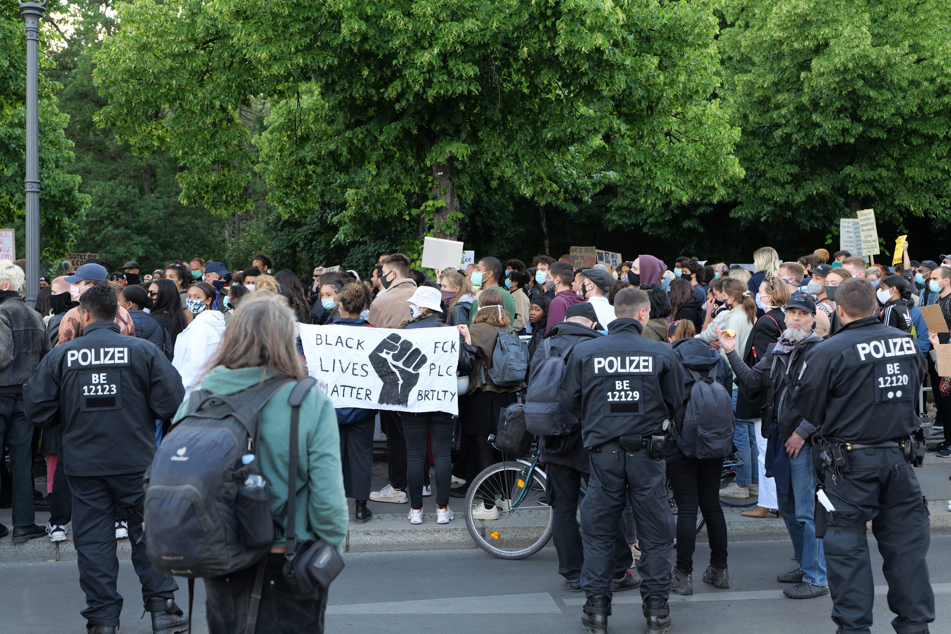 Eine große Gruppe von Menschen mit Schildern und Mützen sowie Taschen steht an einer Straße während einer Black Lives Matter Demonstration in Berlin, mit einem Fahrrad im Vordergrund und Bäumen und einem Pfahl im Hintergrund.
