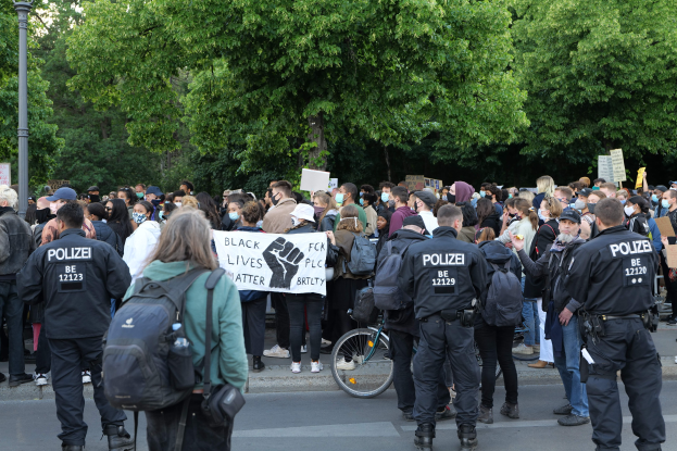 Eine große Gruppe von Menschen mit Schildern und Mützen sowie Taschen steht an einer Straße während einer Black Lives Matter Demonstration in Berlin, mit einem Fahrrad im Vordergrund und Bäumen und einem Pfahl im Hintergrund.