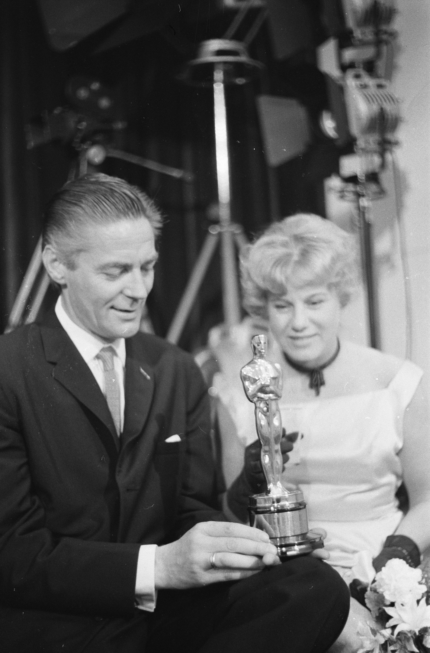 Schwarzes und wei├čes Bild eines formell gekleideten Mannes, der eine Oscar-Statue hält und eine Frau, die einen Blumenstrau├č hält, nebeneinander sitzend mit Hintergrundbeleuchtung.