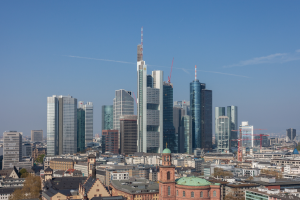 Die Frankfurter Skyline von einem Hügel aus betrachtet, mit einem markanten Wolkenkratzer in der Mitte, verschiedenen Gebäuden und Bäumen im Vordergrund und dem Himmel im Hintergrund.