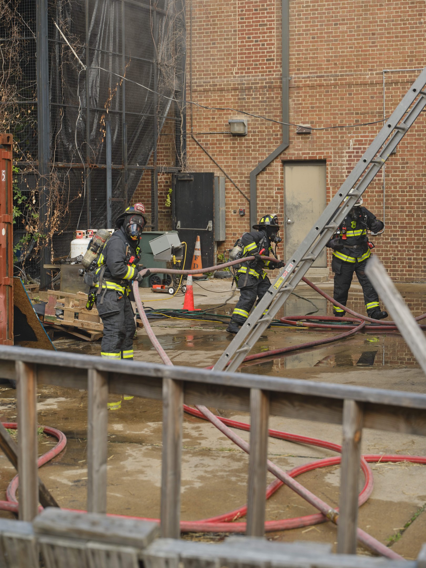 Feuerwehrleute in Helmen arbeiten daran, ein Gebäude Feuer zu löschen, umgeben von Equipment und einem Metallzaun, mit einem Baum und Himmel im Hintergrund.
