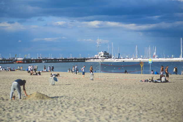 Eine Gruppe von Menschen spielt Volleyball am Strand mit einem Netz, das auf einen Körper von Wasser mit Booten, Pfählen, eine Brücke und einen bewölkten Himmel blickt.