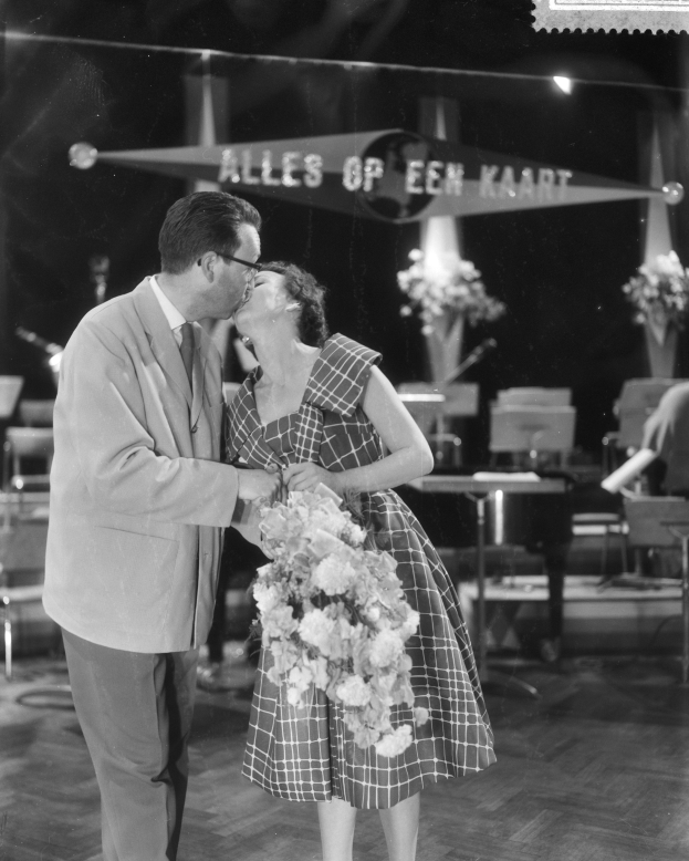 Ein Mann und eine Frau umarmen und küssen sich auf einer Bühne, die Frau hält einen Blumenstrauß, im Hintergrund sind Stühle und Vasen zu sehen.