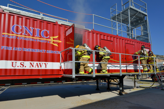 Eine Gruppe von Feuerwehrleuten in Helmen mit Rohren auf einem roten Seecontainer mit Text, Geländern und Stufen, mit einem Fahrzeug auf der Straße, Bäumen und einem bewölkten Himmel im Hintergrund.