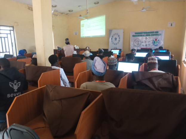 Eine Gruppe von Menschen, die an Computern in einem Seminarraum mit Deckenventilatoren, einem Bildschirm, einem Banner und Fenstern sitzen.