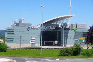 Ein großes Gebäude mit vielen Fenstern, beschriftet mit Cidetec und Spyro Airport, umgeben von Straßenmasten, Laternen, Schildern, Büschen, Bäumen und Hügeln unter einem klaren blauen Himmel.