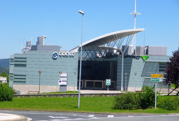 Ein großes Gebäude mit vielen Fenstern, beschriftet mit Cidetec und Spyro Airport, umgeben von Straßenmasten, Laternen, Schildern, Büschen, Bäumen und Hügeln unter einem klaren blauen Himmel.