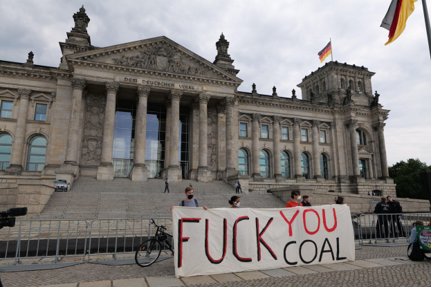 Eine Gruppe von Menschen hält ein Transparent mit der Aufschrift "Fuck You Coal" vor dem Reichstaggebäude in Berlin, Deutschland, mit den architektonischen Details des Gebäudes, einem Fahrrad in der Nähe, Bäumen, einem Fahnenmast und einem bewölkten Himmel.