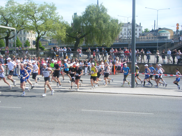 Gruppe von Menschen, die bei einem Marathon auf einer Straße mit Zielpfosten, Band, Absperrungen, Pfosten, Schildern, Brücke, Gebäuden, Bäumen und einem bewölkten Himmel laufen.