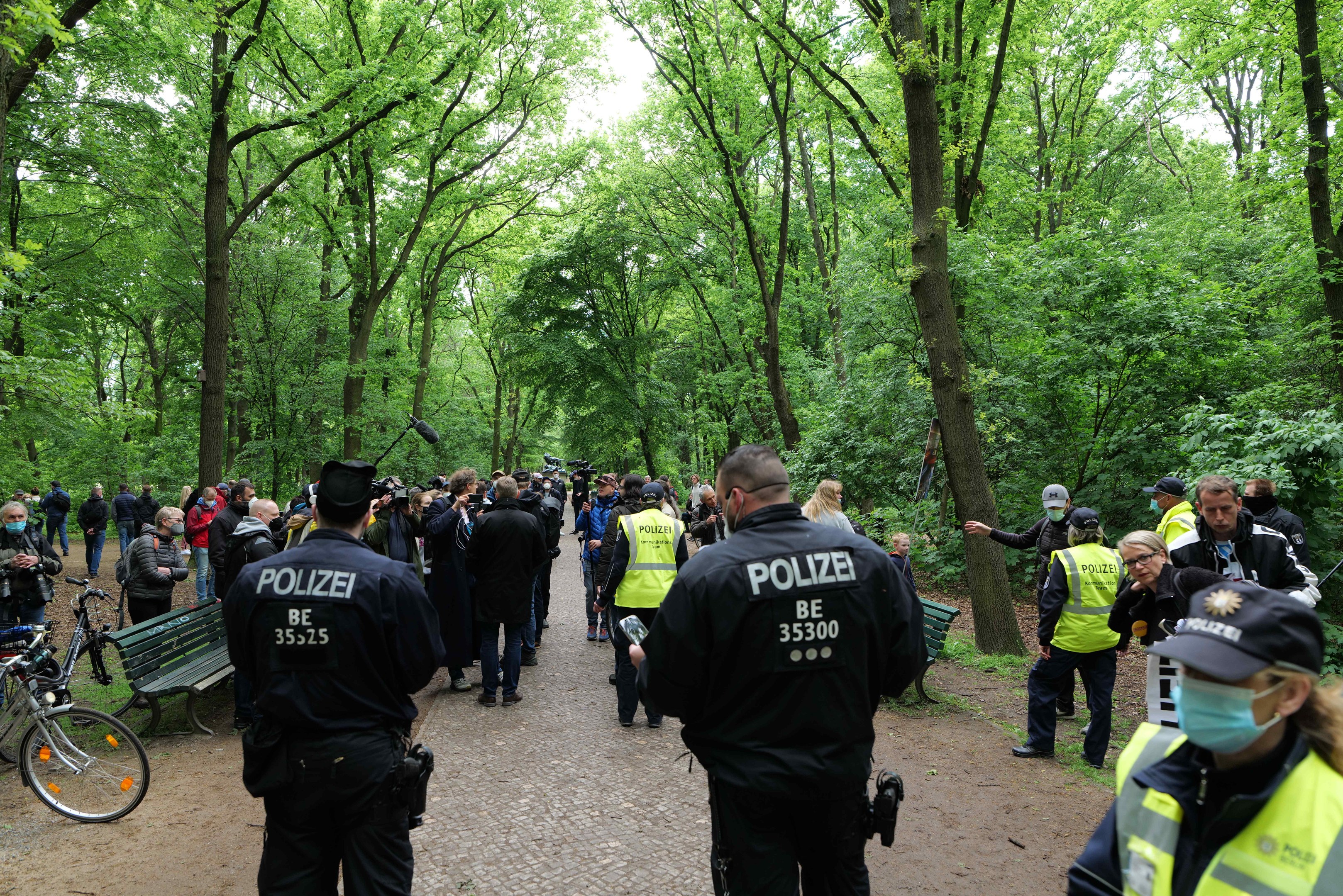 Polizeibeamte in Uniform vor einer Menschenmenge bei einer Outdoor-Veranstaltung, mit Fahrrädern und einer Bank im Vordergrund und Bäumen im Hintergrund.