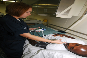 Ein Mann liegt in einem Krankenhausbett, während eine Krankenschwester neben ihm steht und ein elektronisches Gerät hält, mit medizinischer Ausrüstung und an der Wand angebrachten Schränken im Hintergrund.