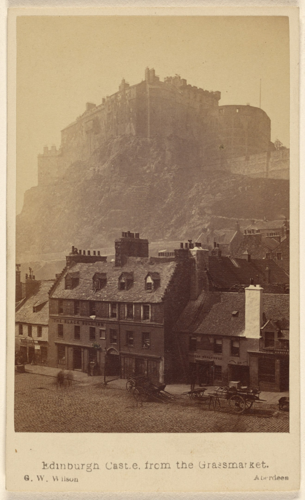 Alte Fotografie des Edinburgh Castle von der Grassmarket aus betrachtet, mit Gebäuden und pferdegezogenen Karren im Vordergrund und einem Hügel im Hintergrund; Text ist unten im Bild sichtbar.