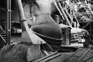 Ein Schwarz-Weiß-Foto einer großen Maschine in einer Fabrik, umgeben von verschiedenen Maschinen, Polen, Treppen, Geländern und anderen Gegenständen, mit Eisenbahnschienen unten.